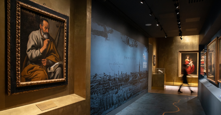 View of a corridor inside The Spanish Art Gallery at The Auckland Project with paintings on the wall and a woman walking past in the background. 